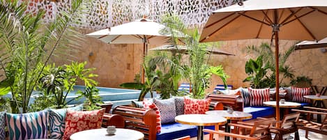 An outdoor patio area with wooden tables and chairs, covered by umbrellas, and surrounded by plants and a decorative wall.