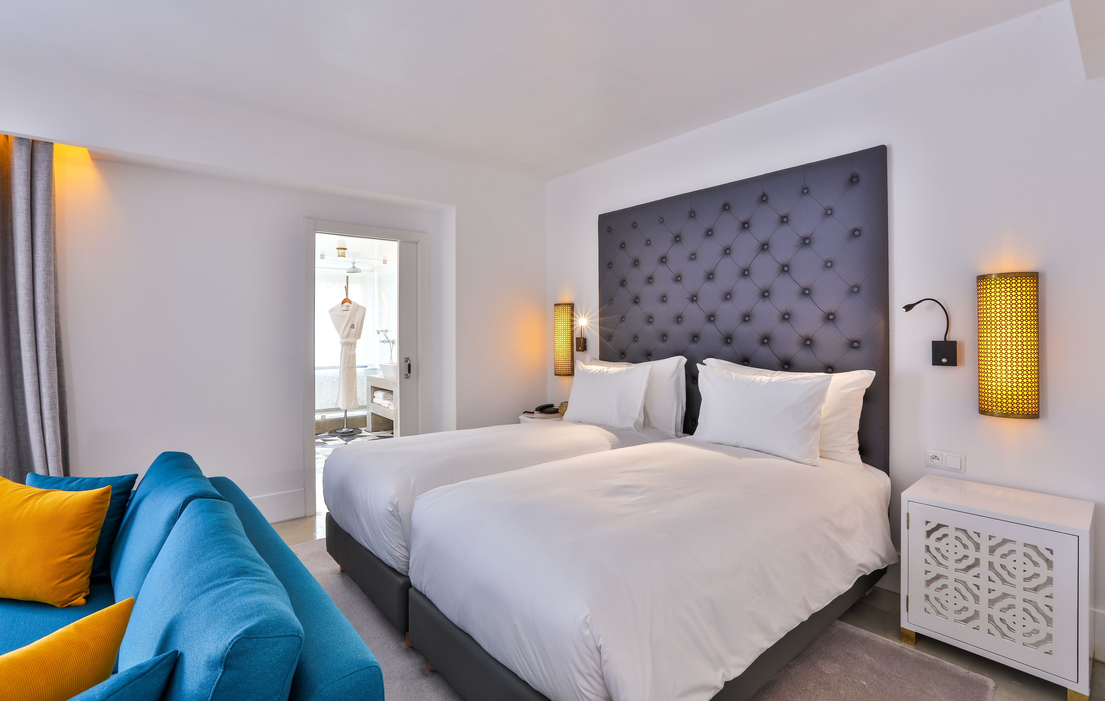 A hotel room with two beds, a blue sofa, and a white nightstand with a lamp.