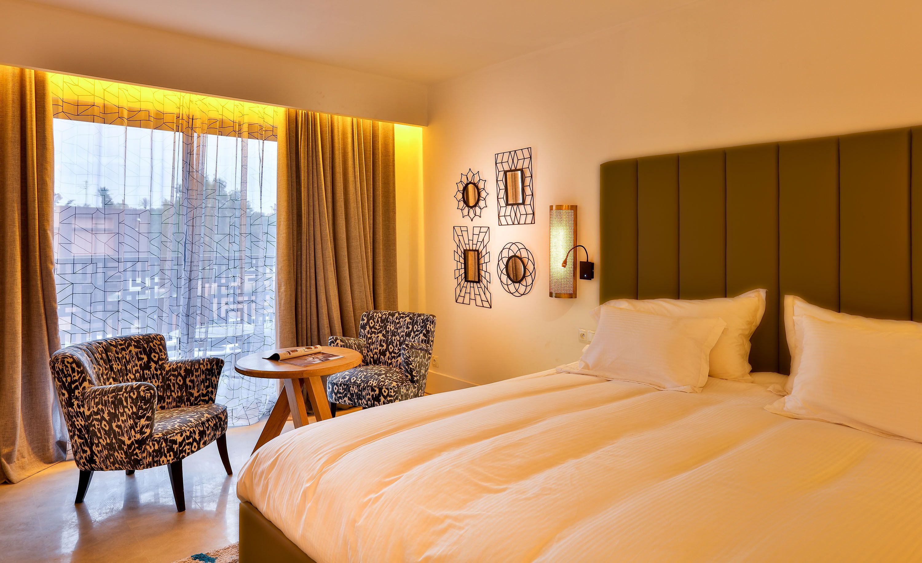 A hotel room with a bed, two patterned armchairs, a small round table, and a view of buildings through sheer curtains.