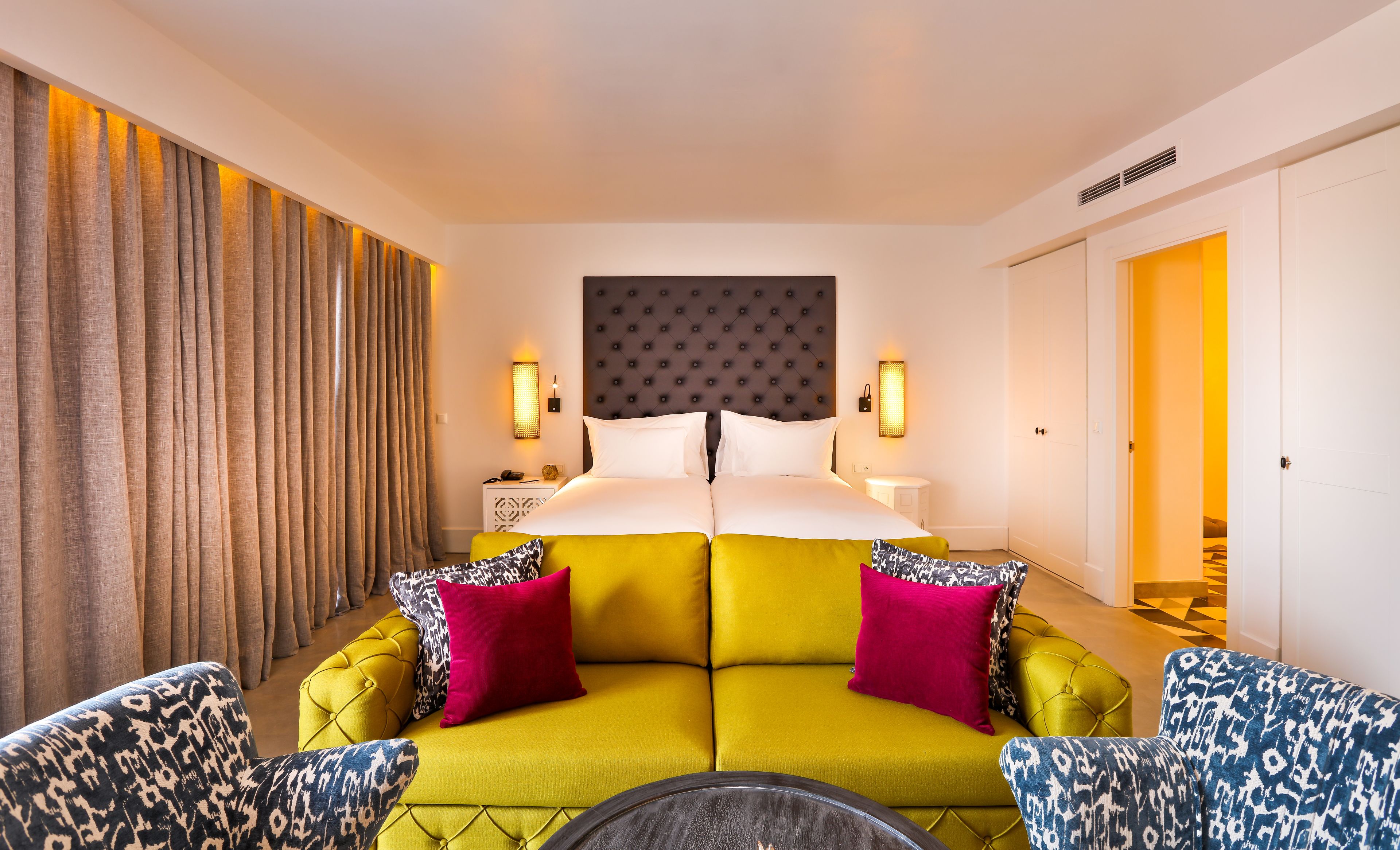 Habitación de hotel con una cama grande, un sofá, dos sillones, una mesita de noche y una lámpara de pared.