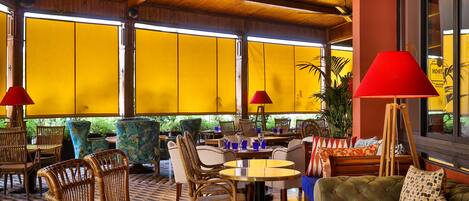 A spacious lounge area with wicker furniture, a ceiling fan, and large windows with yellow blinds.
