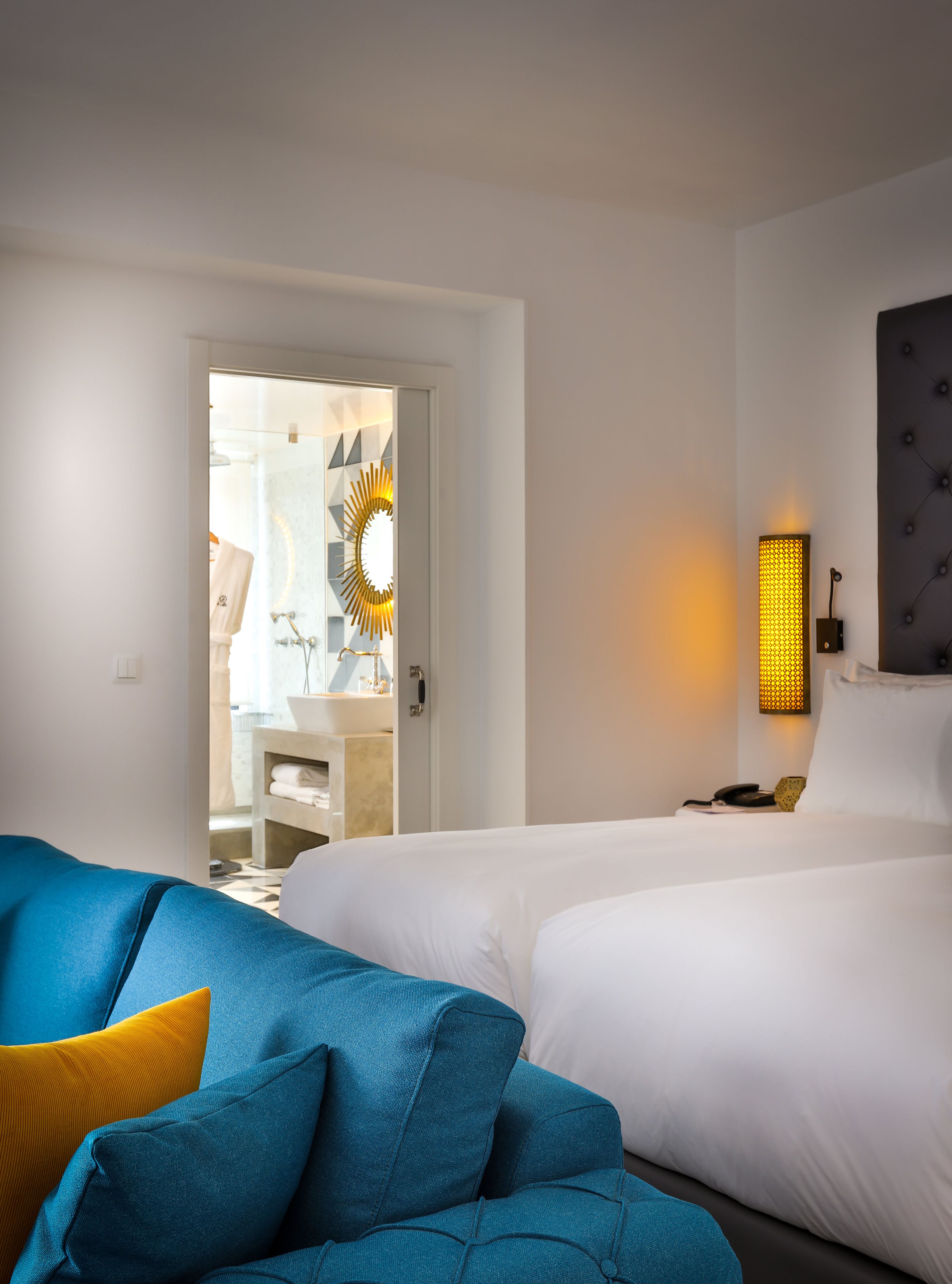 Habitación de hotel con dos camas, baño con espejo y toallero, y sofá azul con un cojín amarillo.