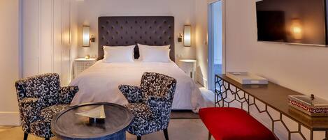 Habitación de hotel con una cama grande, dos sillones, una mesita redonda, un banco rojo, un televisor y una mesita de noche con lámpara.