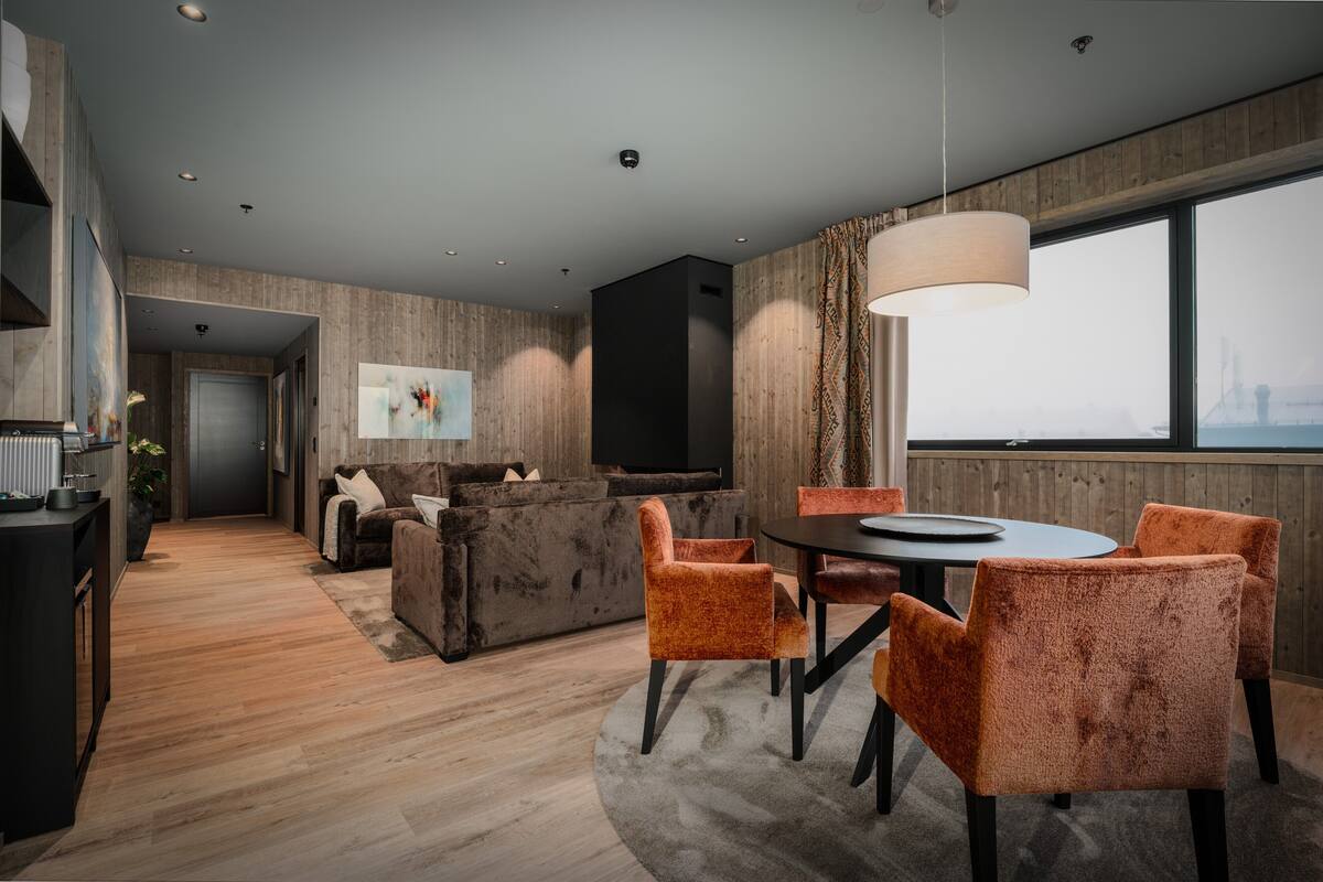 Un salon moderne avec un canapé foncé, une table basse et des fauteuils orange. On y trouve également un coin repas avec une table ronde et des chaises. La pièce est dotée d’un parquet et d’une grande fenêtre offrant une vue sur le paysage urbain.