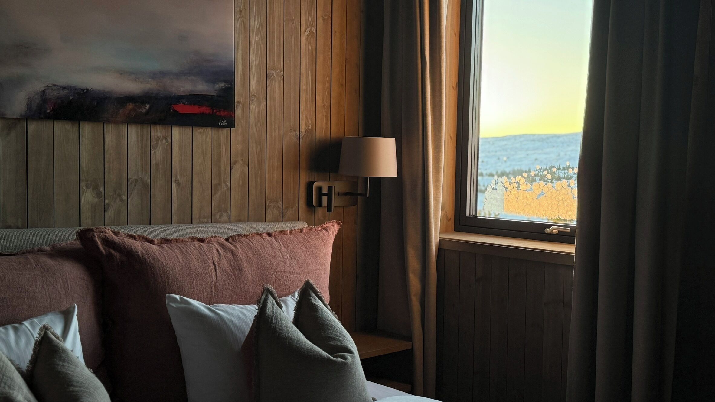 A hotel room with a wooden wall, a bed with multiple pillows, a nightstand, a lamp, and a window with a view of the sea.