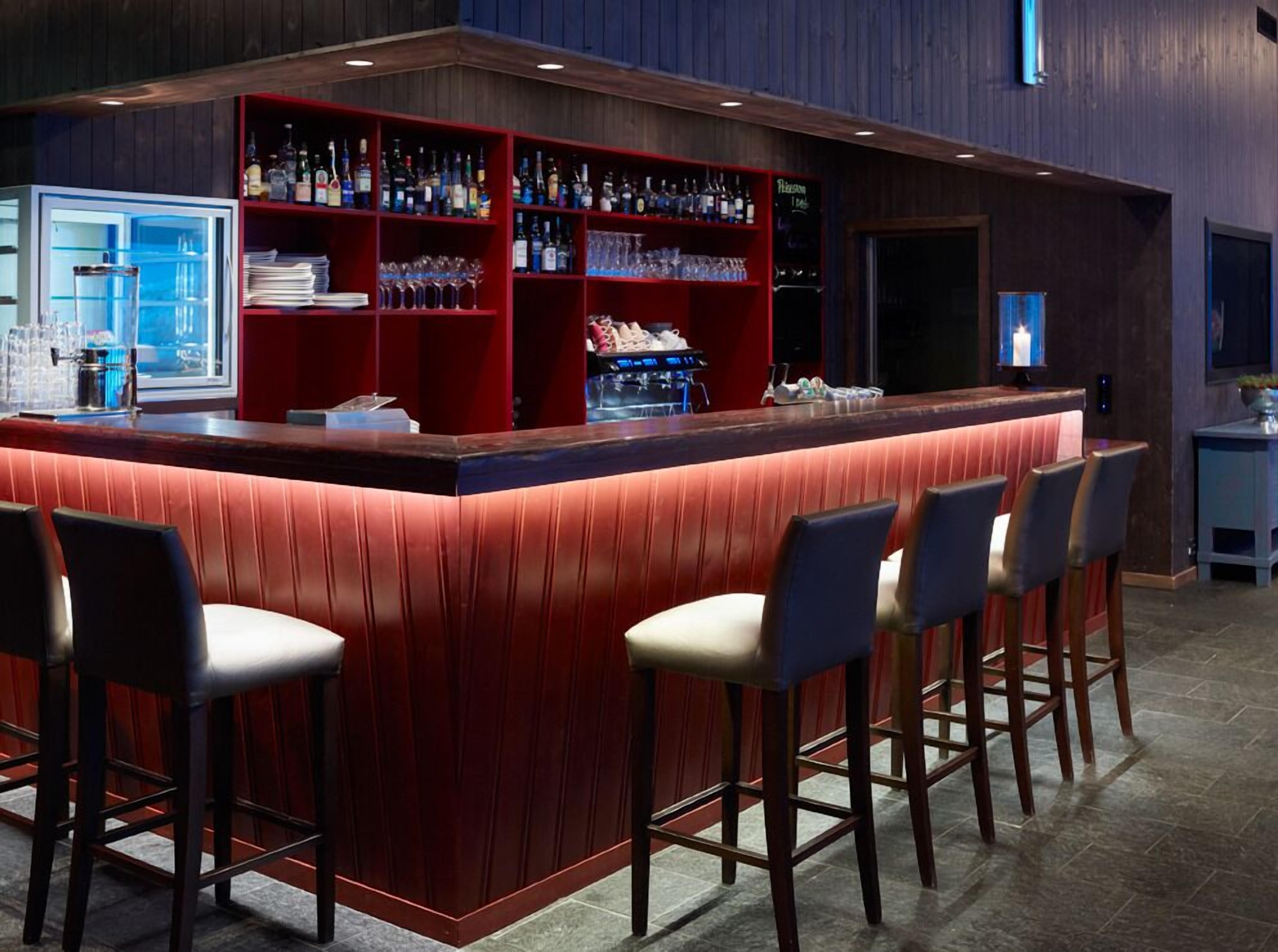 A bar area with a red counter, shelves with bottles, glasses, and a refrigerator. There are four bar stools in front.