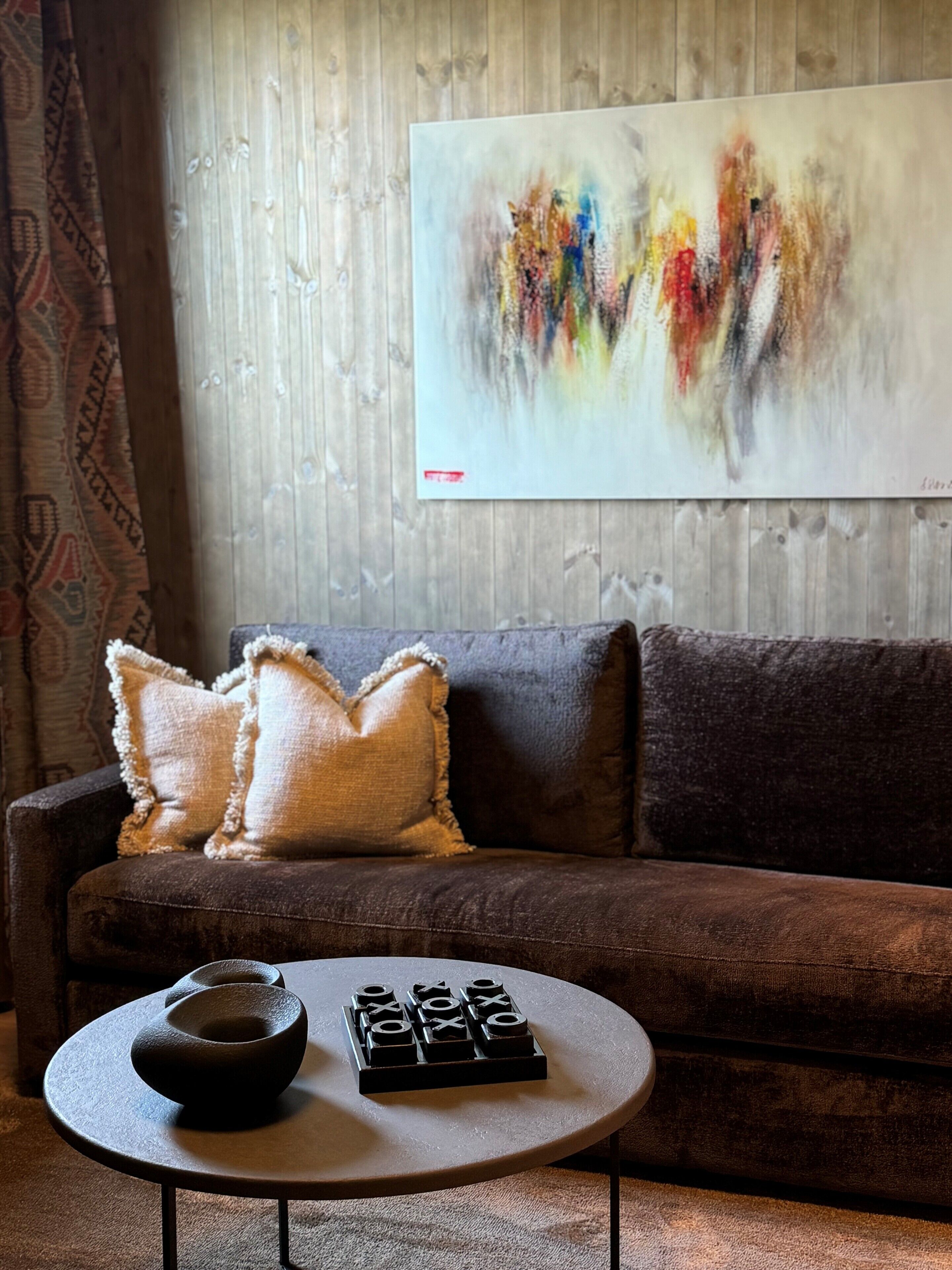 Una sala de estar moderna con un sofá marrón, dos almohadas con textura, una mesa de centro redonda con un cuenco y un juego de mesa, y un cuadro abstracto colorido en la pared.