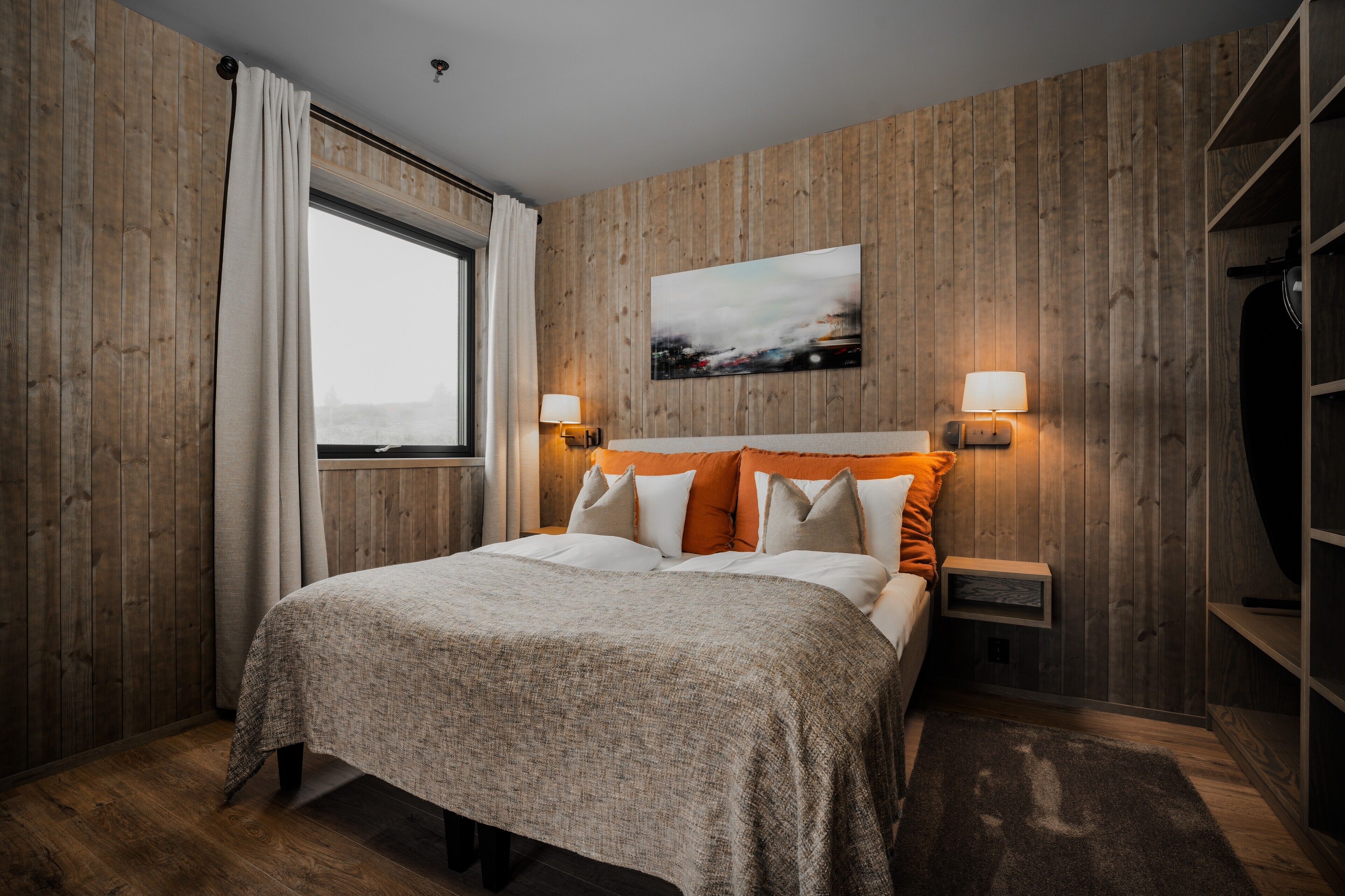 Un dormitorio con panel de pared de madera, una cama grande con cabecera naranja, dos lámparas de noche y un ventanal con cortinas.