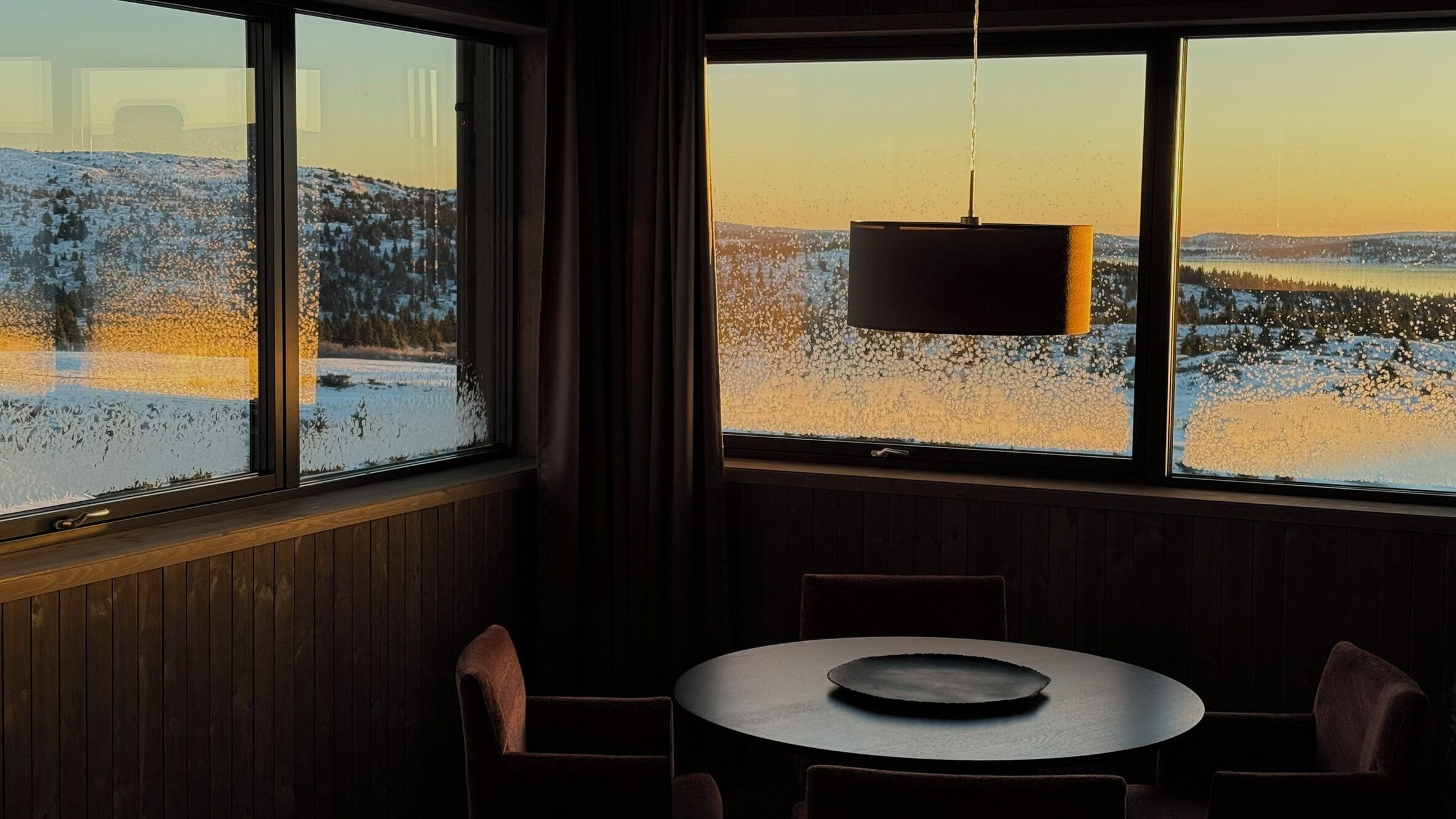 Ein Zimmer mit einem großen Fenster mit Blick auf eine verschneite Landschaft, einem runden Tisch mit Stühlen und einer Hängeleuchte.