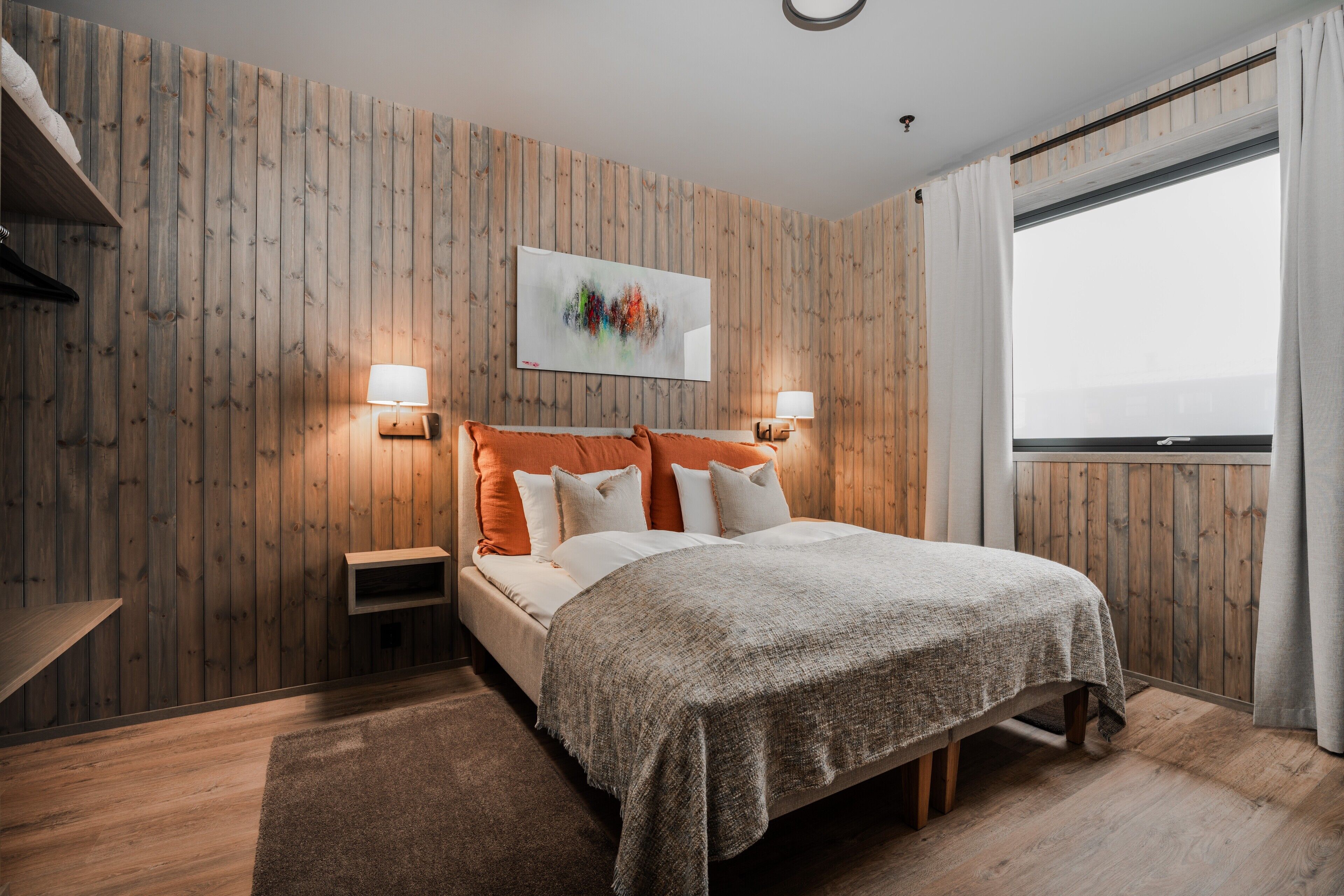 Ein Schlafzimmer mit einer Holzvertäfelung, einem Bett mit weißer und orangefarbener Bettwäsche, zwei Nachttischlampen und einem Fenster mit Vorhängen.