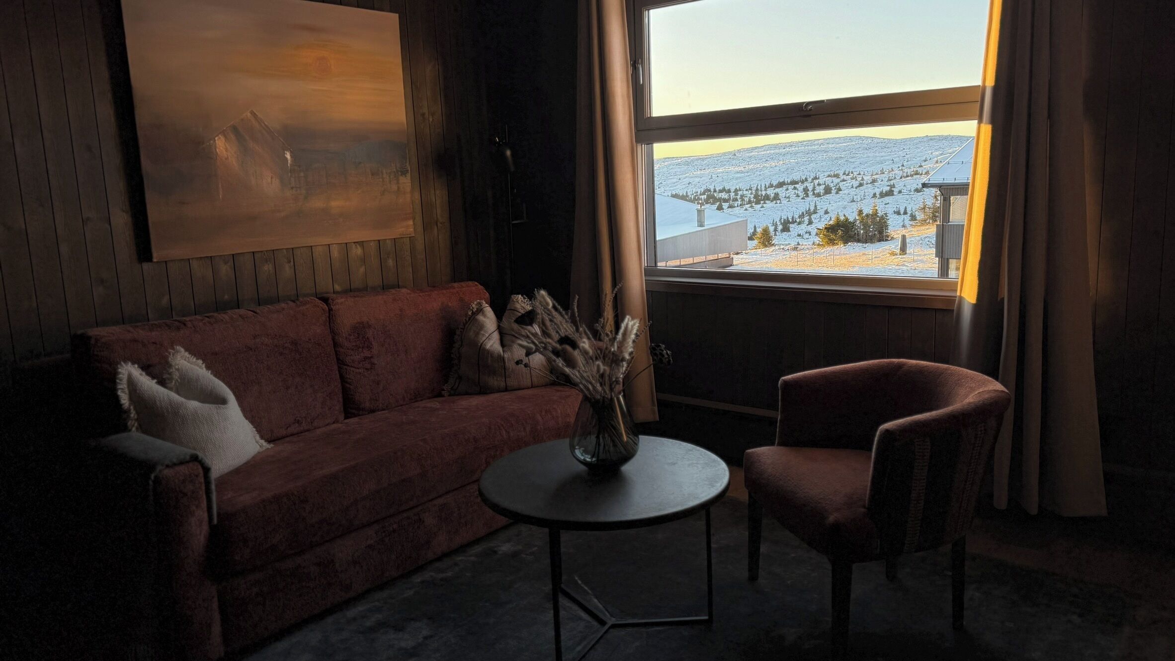 Una habitación acogedora con un sofá rojo, una mesita redonda y una silla. Un cuadro adorna la pared, y hay una ventana con vistas a un paisaje nevado.