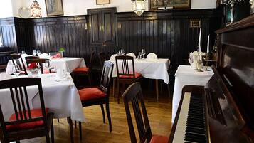 A dining area with tables set for two, wooden chairs, a piano, and wall-mounted chandeliers.
