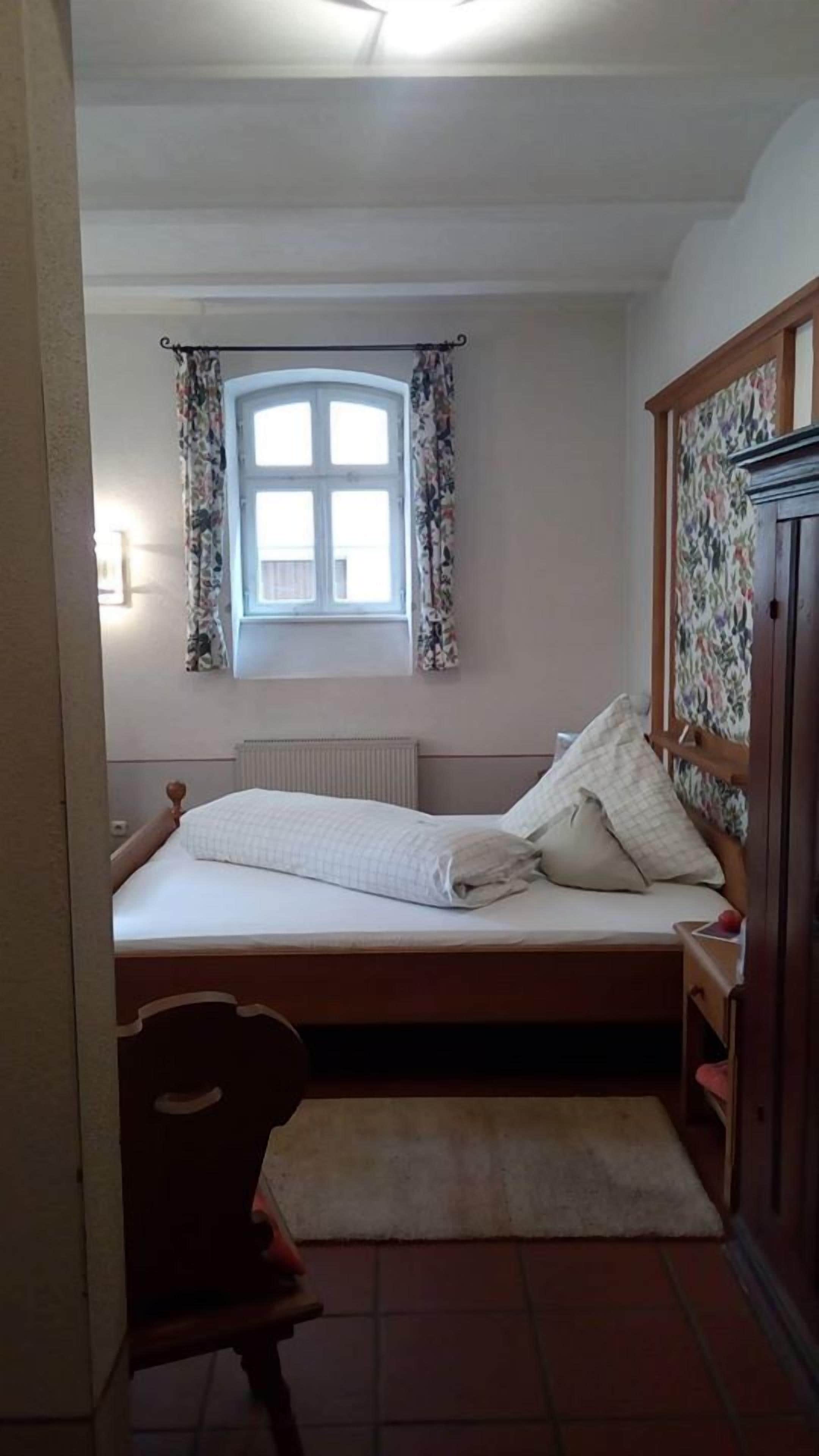 A bedroom with a bed, a window with floral curtains, a radiator, and a wooden wardrobe.