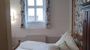 A bedroom with a bed, a window with floral curtains, a radiator, and a wooden wardrobe.