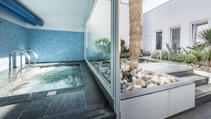 Ein moderner Poolbereich im Innenbereich mit großen Glaswänden, einer Wand aus blauen Mosaikfliesen und einem Steinweg, der zu einer Palme führt.