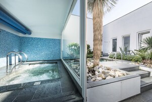 A modern indoor pool area with clear glass walls, a blue mosaic tile wall, and a stone pathway leading to a palm tree.