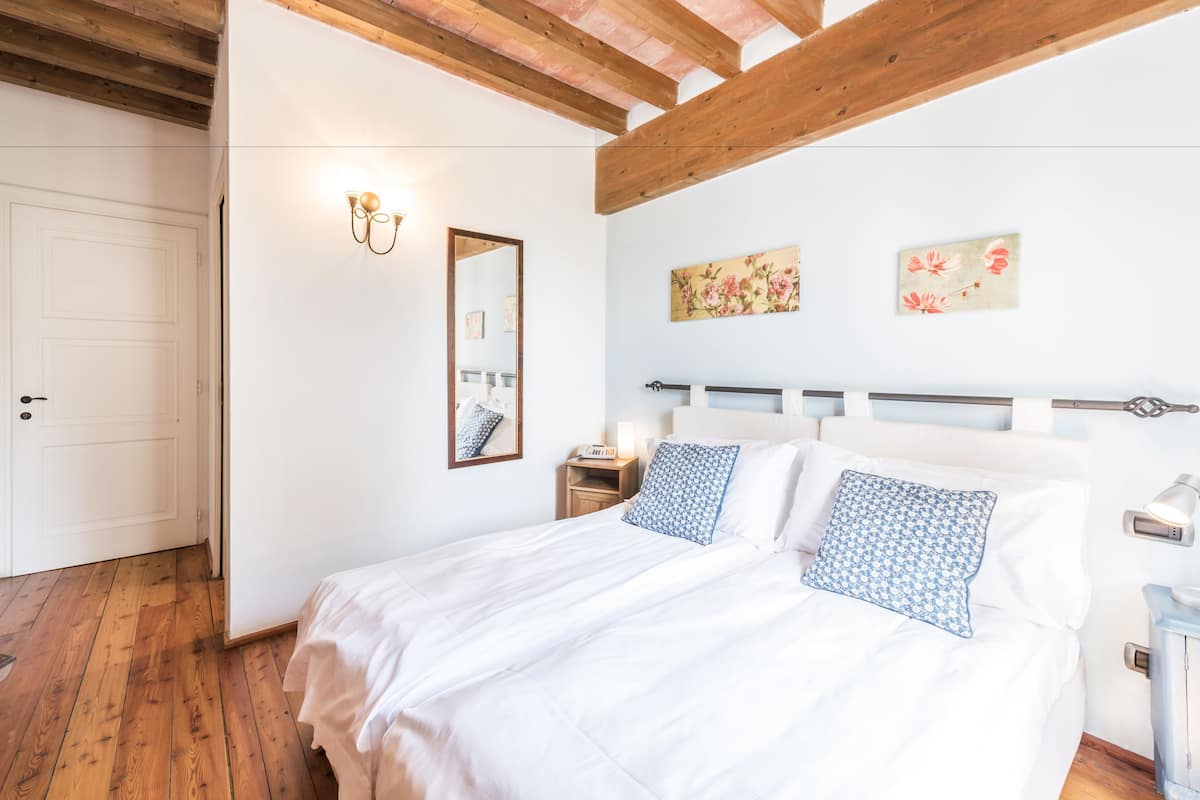 Ein Schlafzimmer mit Holzboden, einem Bett mit weißer Bettwäsche und gemusterten Kissen, einem Nachttisch mit Lampe und Wandbildern.