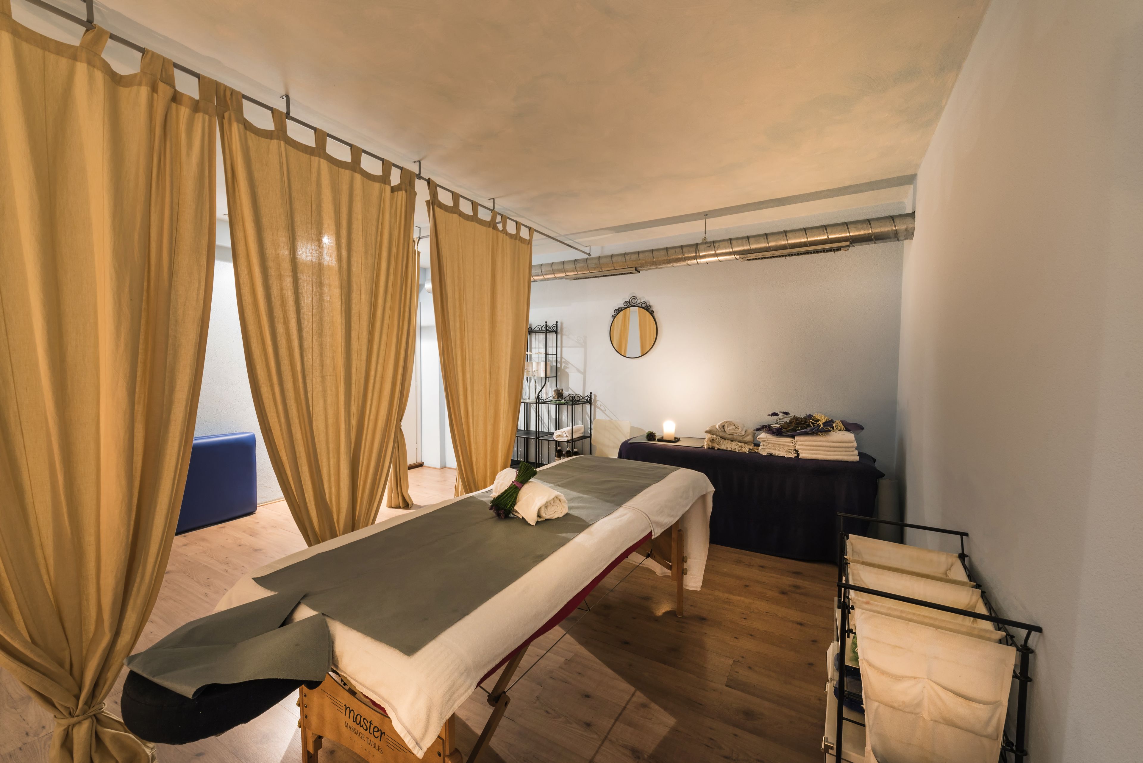 A room with a massage table, curtains, a mirror, a small table with towels, and a rack with towels.