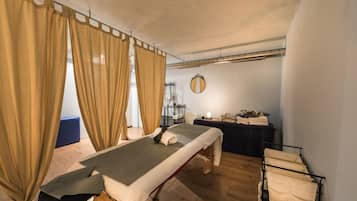 A room with a massage table, curtains, a mirror, a small table with towels, and a rack with towels.