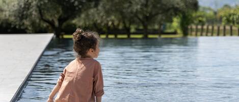 A child sitting by a calm water body, with trees and a clear sky in the background.