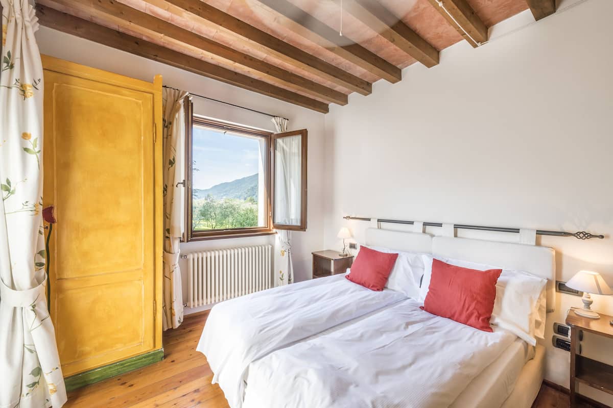 Ein Schlafzimmer mit Holzboden, einem Bett mit weißer und roter Bettwäsche, einem Fenster mit Blick auf Grünflächen und einer Heizung.