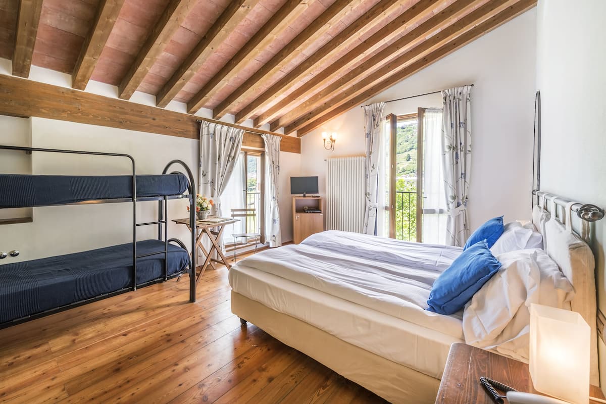 Ein Zimmer mit Etagenbett, einem Einzelbett, Holzboden, einem Fenster mit Vorhängen und einem kleinen Tisch mit Stuhl.