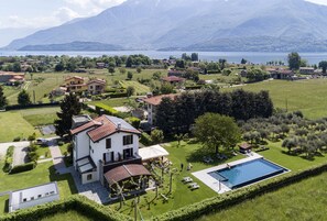 A large house with a swimming pool, surrounded by lush greenery and a lake view.
