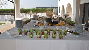 Una mesa de buffet con diversos alimentos y bebidas, dispuesta al aire libre con manteles blancos y sillas.