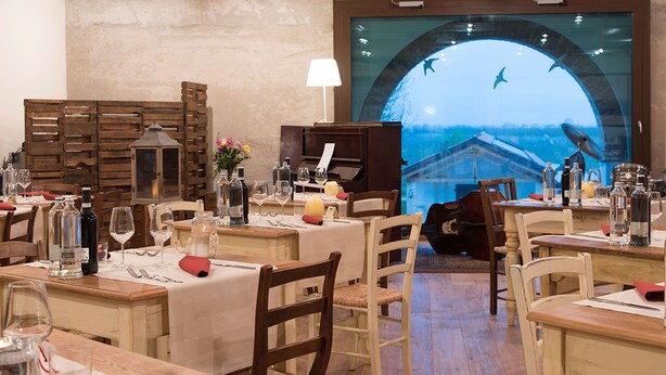 Un restaurante con mobiliario de madera, manteles blancos y un amplio ventanal con vistas a un edificio y pájaros.