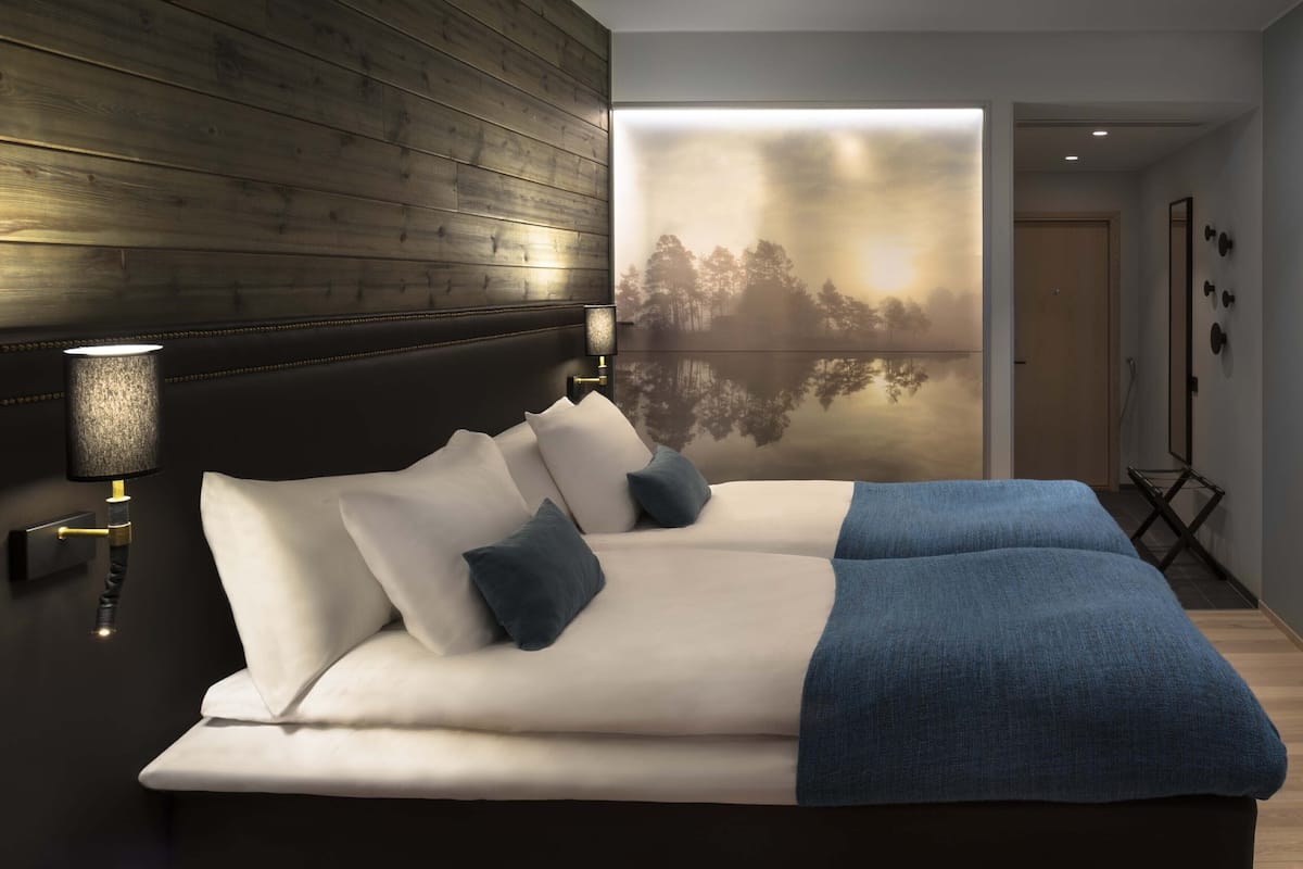 Quarto com cama, mesa de cabeceira, luminária na parede e grande janela com vista para árvores.
