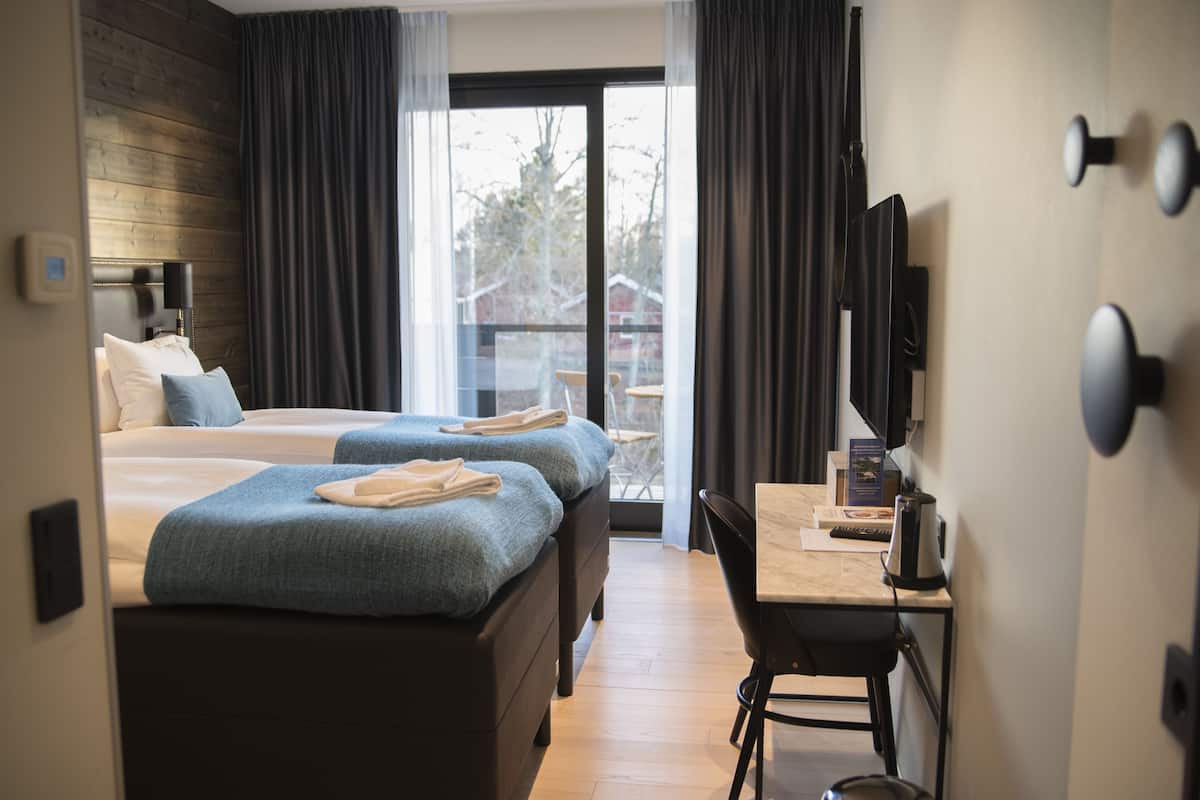 Quarto de hotel com duas camas, uma escrivaninha, uma cadeira, televisão e vista para o exterior.