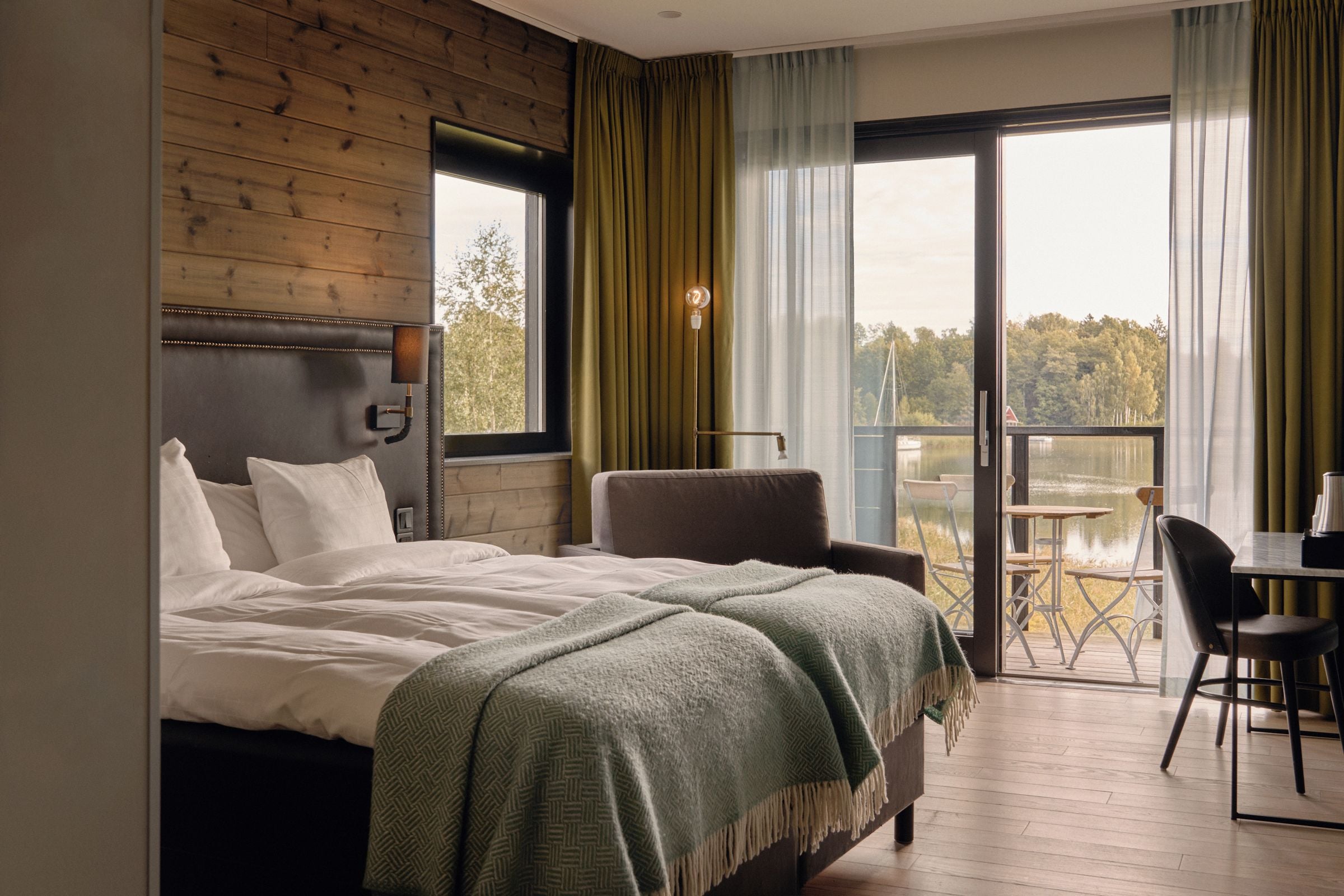 A bedroom with a bed, a chair, a table, and a balcony with a view of a lake.