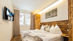 A bedroom with a wooden headboard, a bed with white bedding, a mounted TV, and a window with curtains. - Living Schönwies (Zell am See)