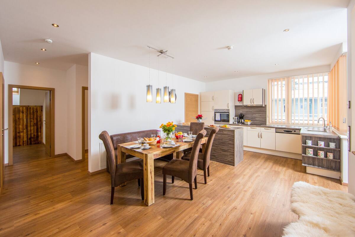 A modern kitchen with wooden cabinets, a dining area with a wooden table and chairs, and a cozy living space with a white fur rug.