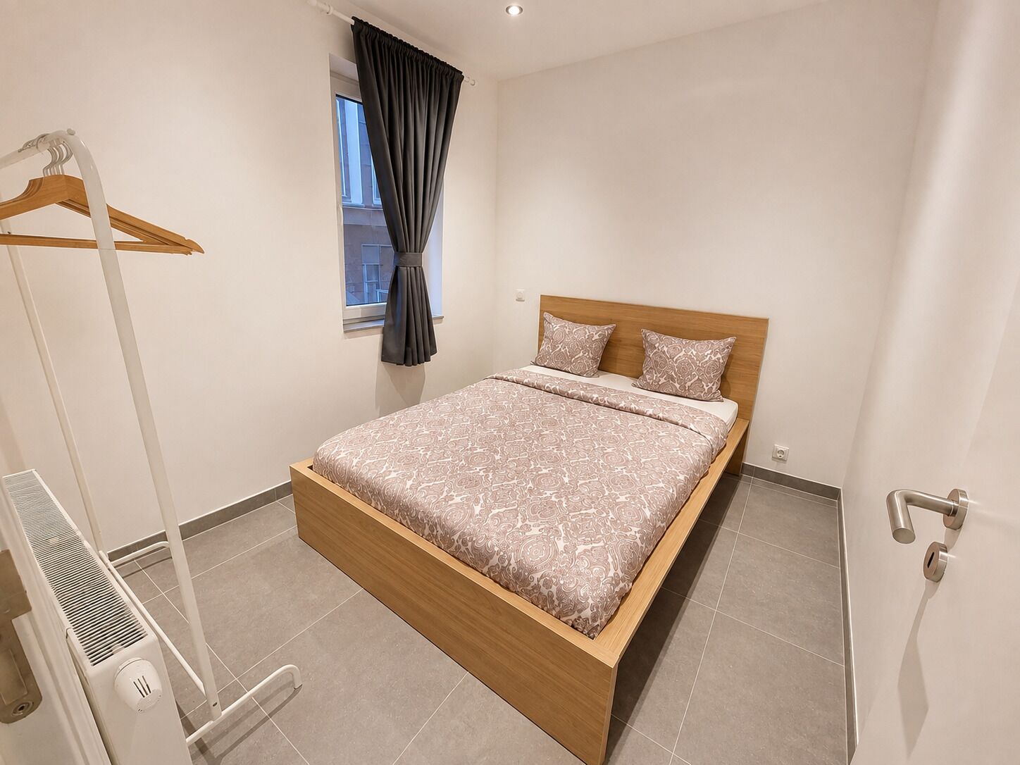 Ein Schlafzimmer mit einem hölzernen Bett, einem fenster mit Vorhängen, einem Heizkörper und einer Tür mit Griff.