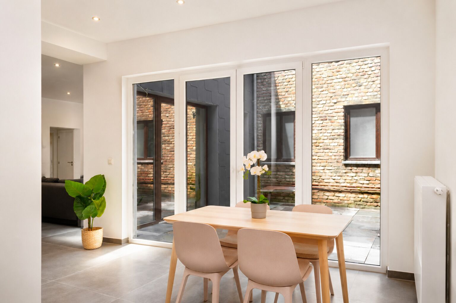 A modern dining area with a table and chairs, a potted plant, and a view through large glass doors to an outdoor brick wall.