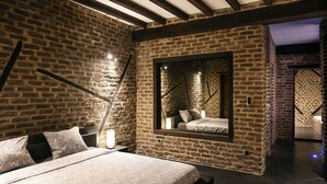 A bedroom with a brick wall, a bed with patterned bedding, a nightstand with a lamp, and a window with a view of another room. - Appart-Hotel Léopold Liège Centre (Liege)