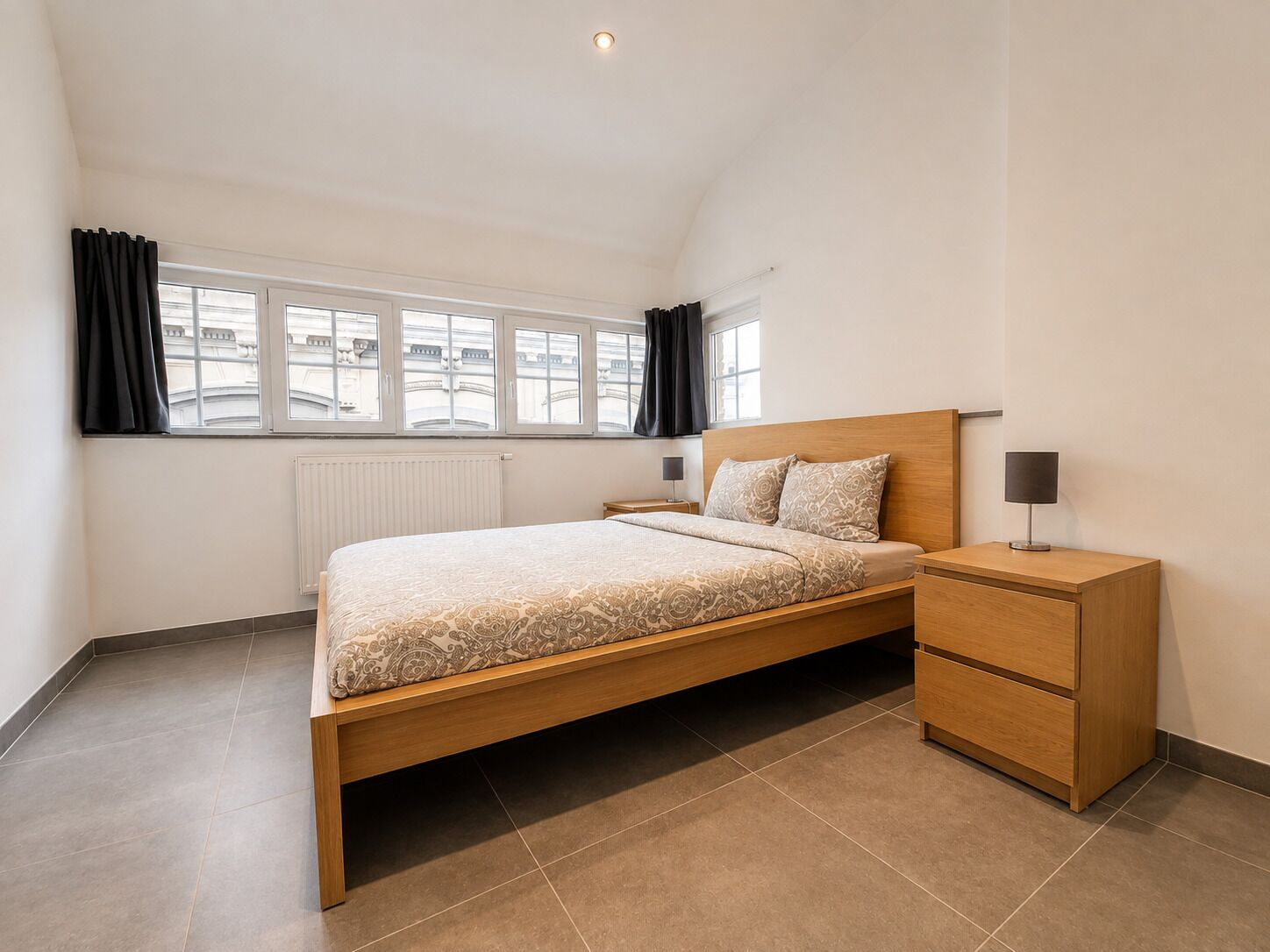 Quarto moderno com cama de madeira, mesa de cabeceira com abajur e vista para edifícios pela janela.