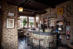 Uma área de bar com paredes de pedra, vigas de madeira e banquetas. Há plantas em vasos e uma variedade de garrafas nas prateleiras.
