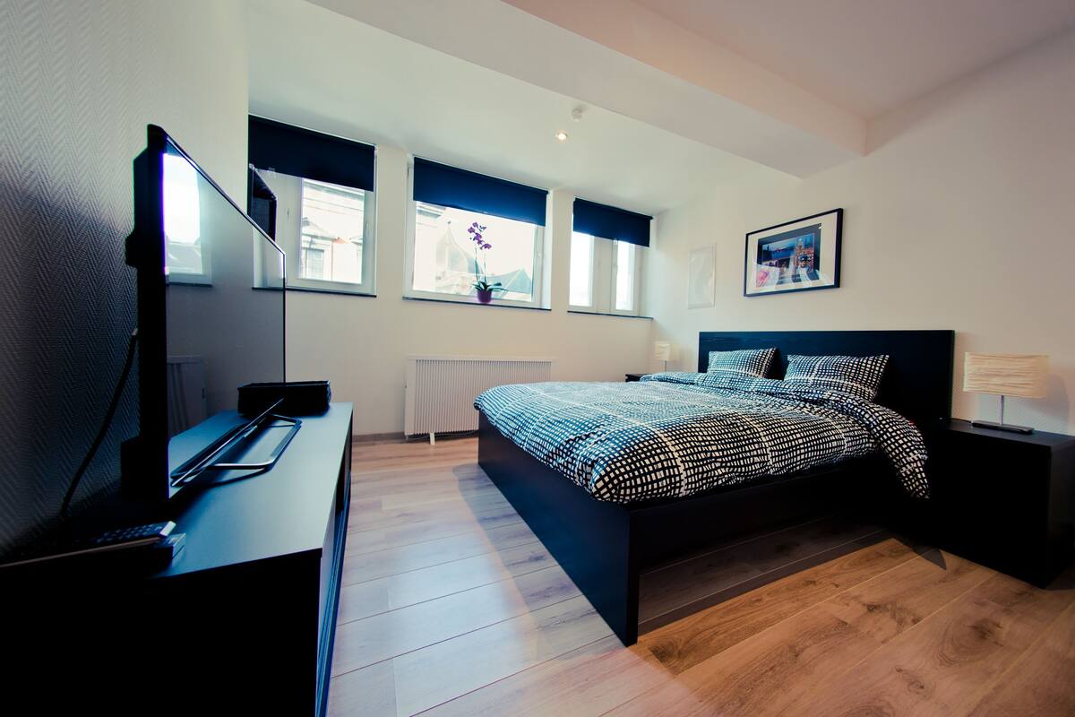 A modern bedroom with a large bed, a desk with a computer, and a window with a view of a building.