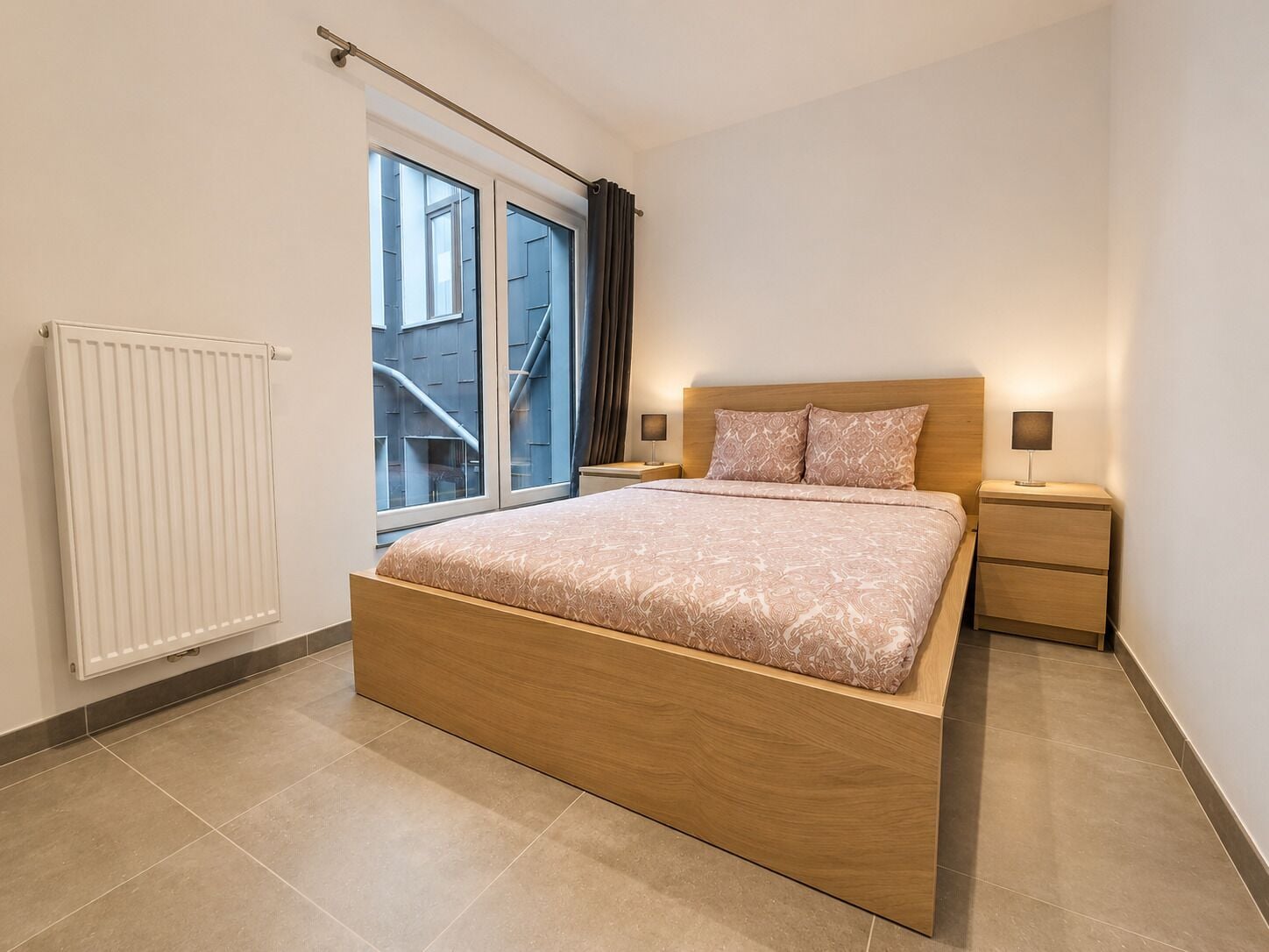 A modern bedroom with a wooden bed, a window with curtains, a radiator, and bedside lamps.