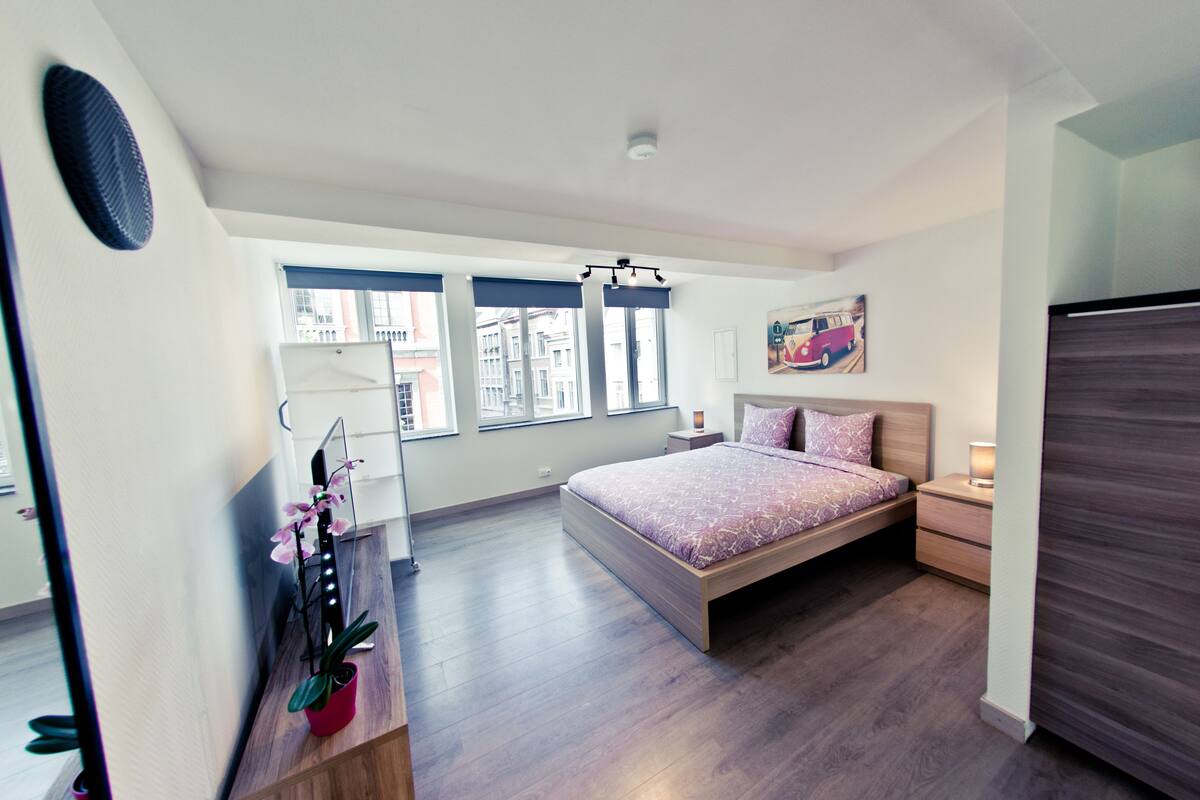 A modern bedroom with a large bed, a nightstand, a bedside table, a potted plant, and a window with a view of buildings.