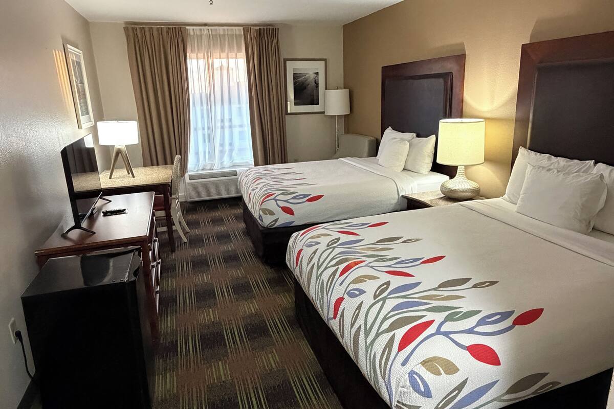 Quarto de hotel com duas camas, uma escrivaninha, uma cadeira, um abajur e uma janela com cortinas.