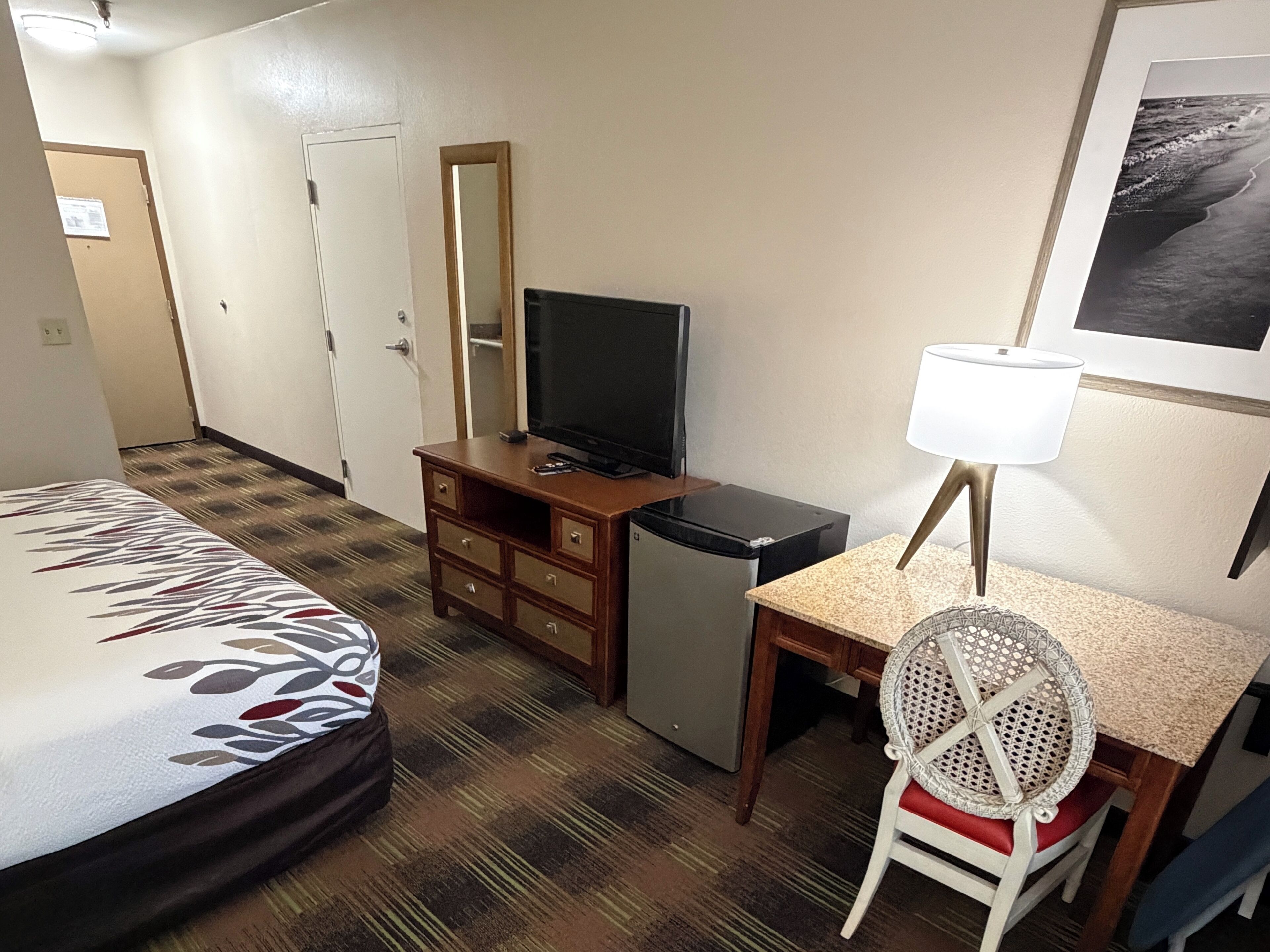 A hotel room with a bed, a desk with a chair, a television, a refrigerator, a mirror, and a framed picture on the wall.