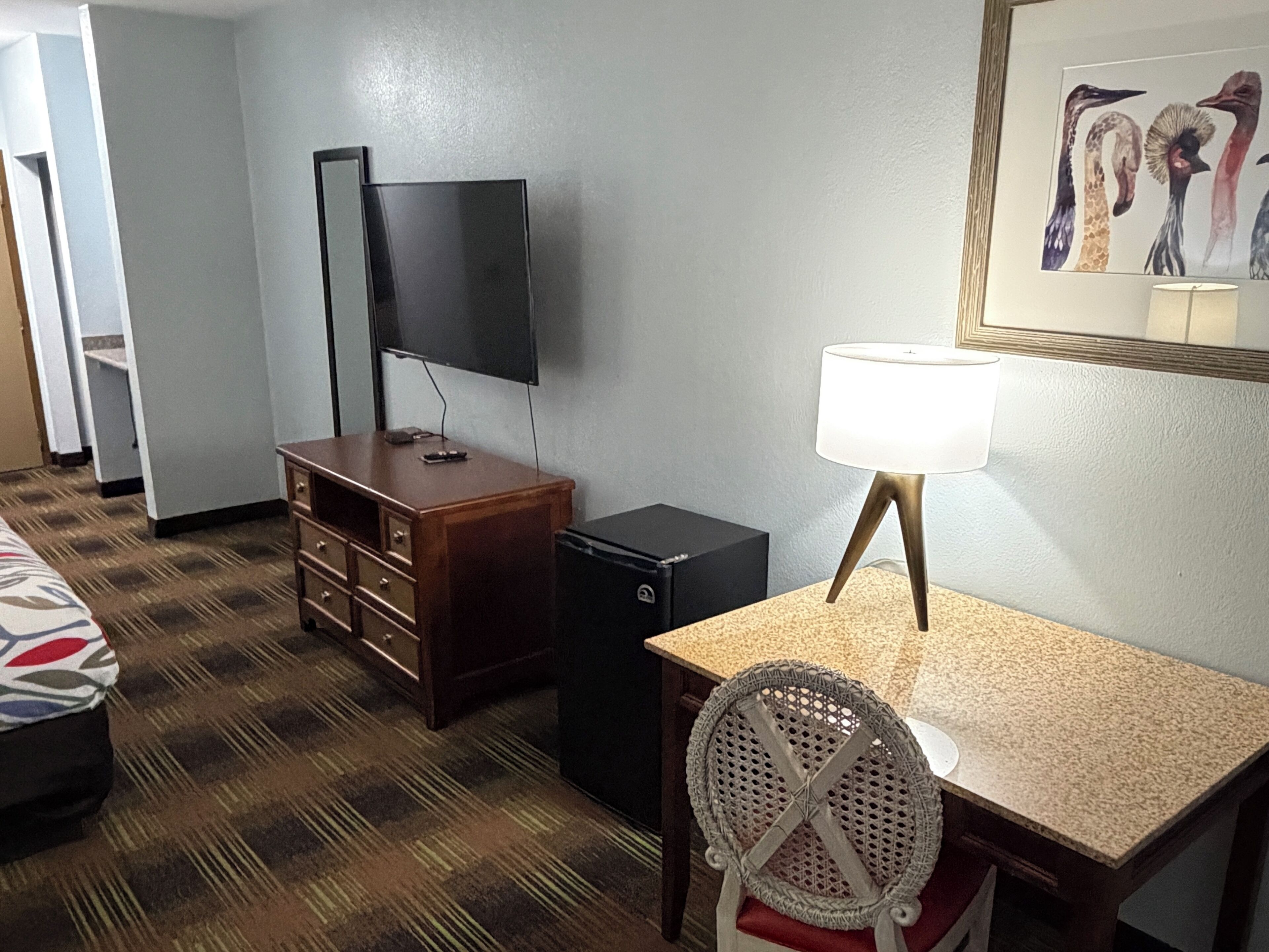 A hotel room with a bed, a desk with a lamp, a TV, a dresser, and a small black speaker.