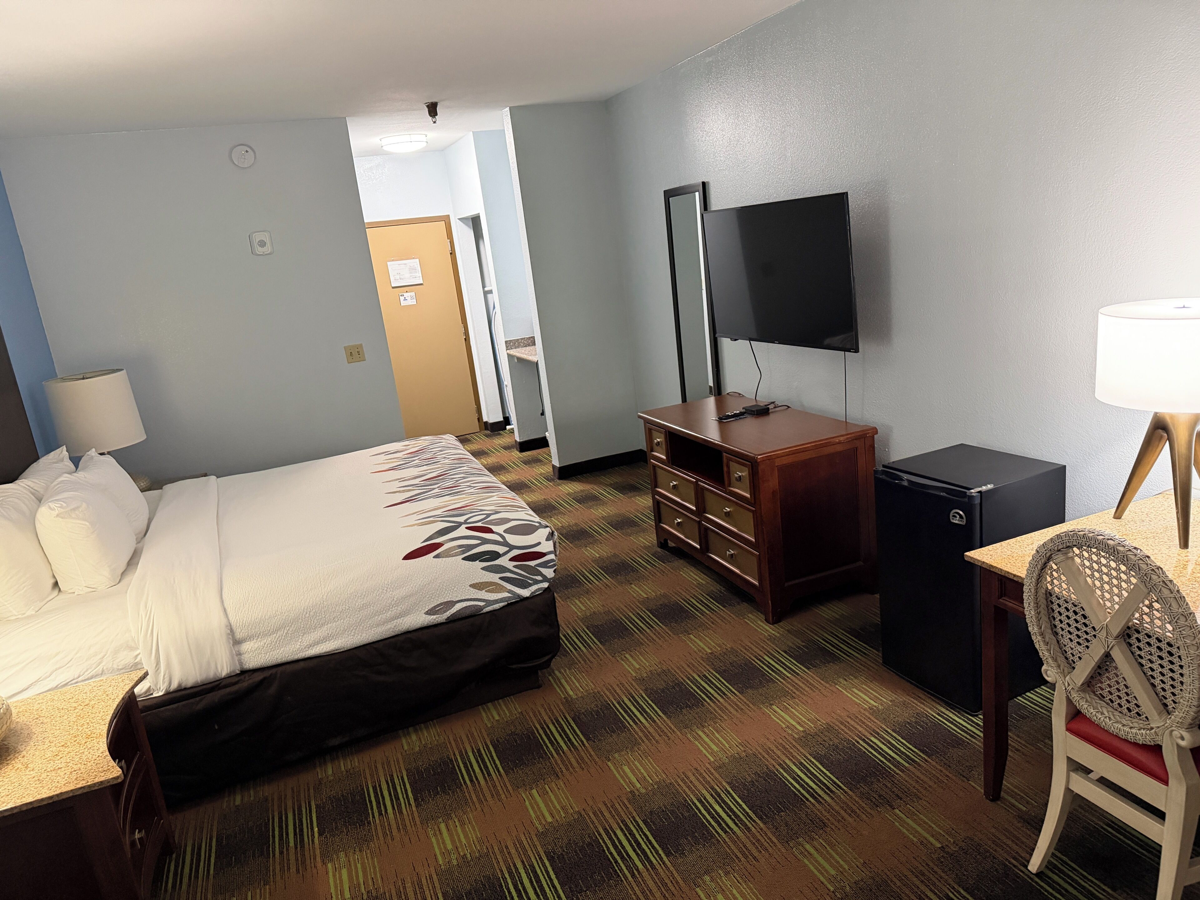 A hotel room with a bed, a desk, a chair, a TV, a small cabinet, and a door leading to a bathroom.