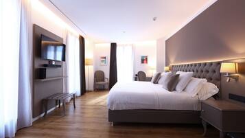A modern hotel room with a large bed, bedside tables, a flat-screen TV mounted on the wall, and a seating area with a bench and chairs.