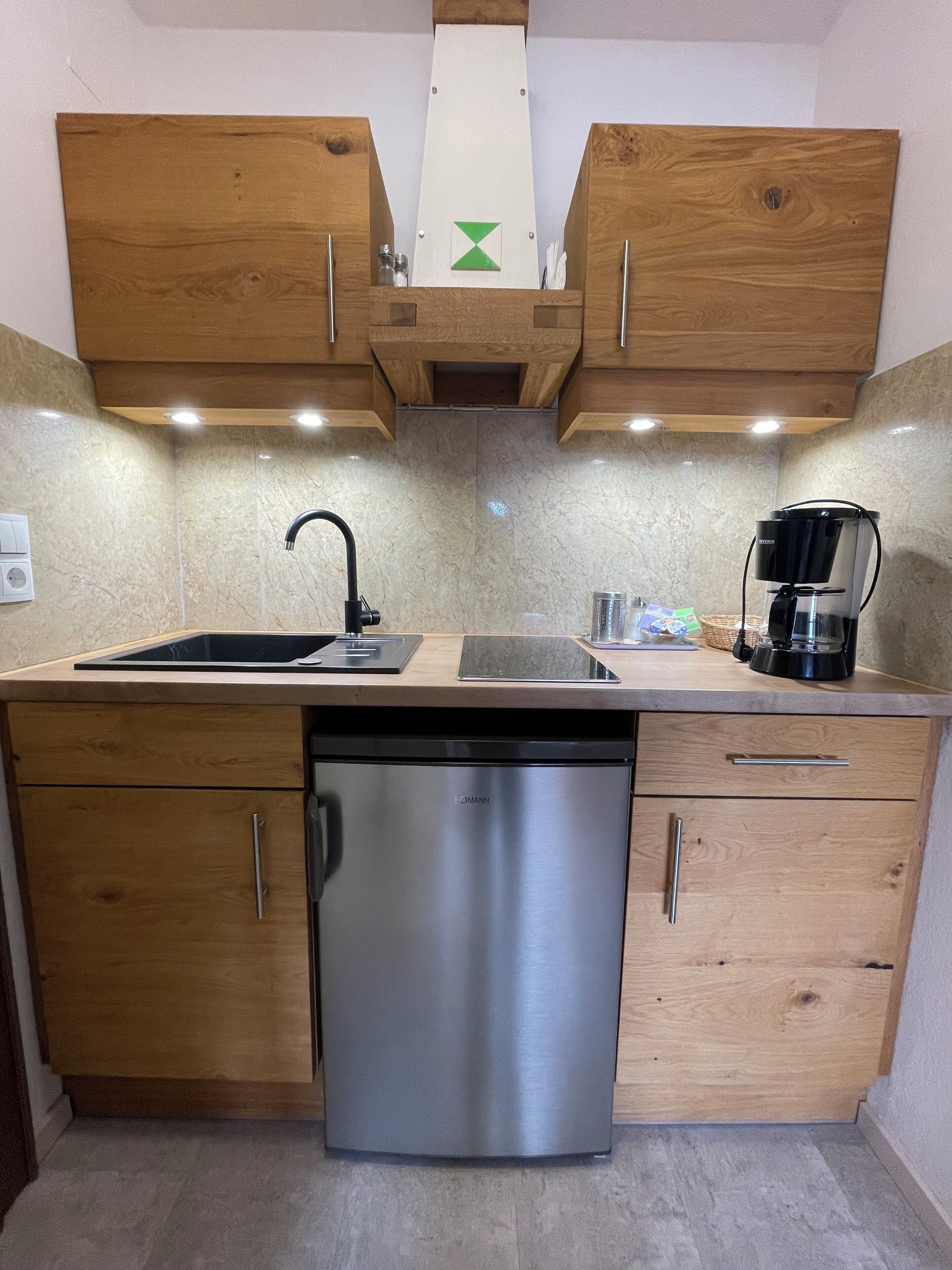 A compact kitchen with wooden cabinets, a stainless steel dishwasher, a black coffee maker, and a marble backsplash.