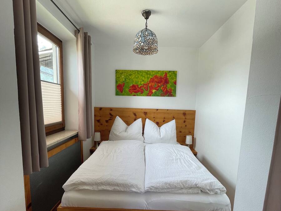 Un dormitorio con cama de madera, una ventana con cortinas, una lámpara de pared y un cuadro con motivos florales.