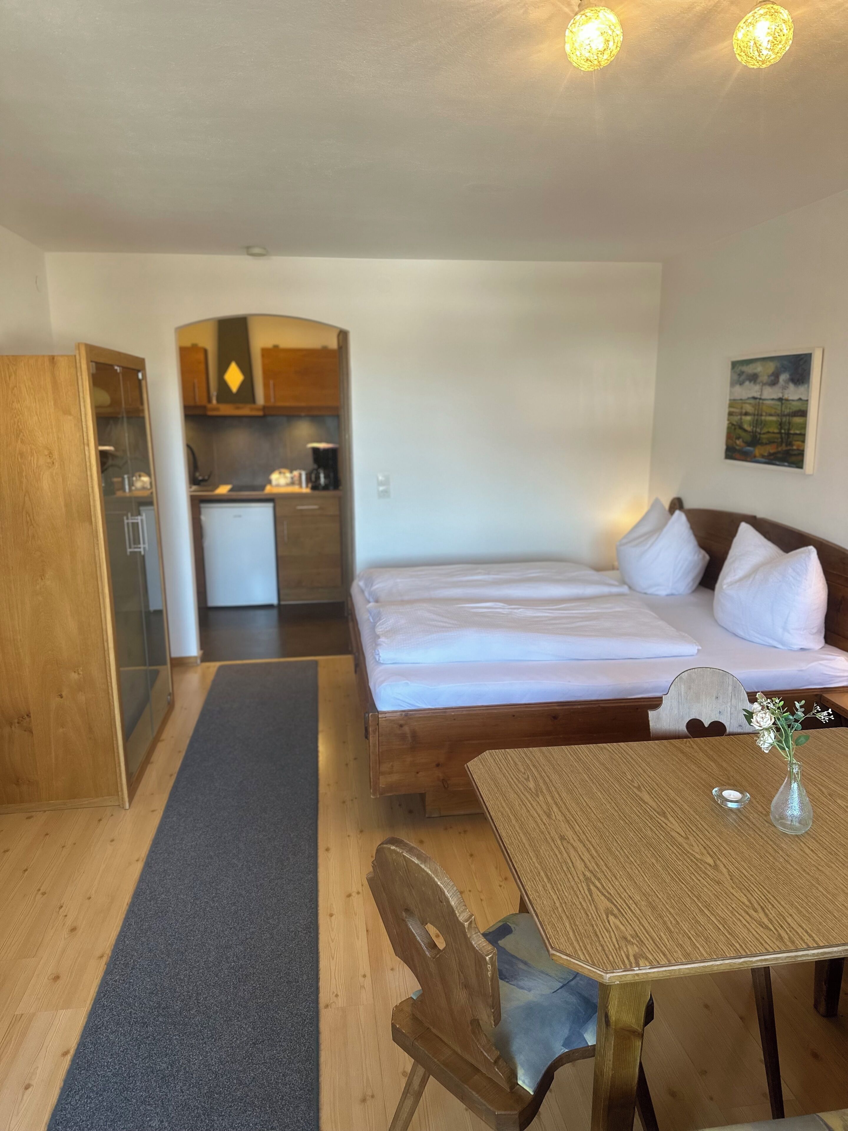 A room with a bed, a wooden table, a chair, a small kitchen area, and a painting on the wall.